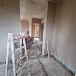 Wnętrze w trakcie prac wykończeniowych: ściany pokryte świeżym tynkiem, widoczna drabina malarska i agregat tynkarski z przewodami na podłodze.