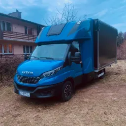Niebieski samochód dostawczy Iveco z plandeką, zaparkowany na trawiastym terenie przed domem w pochmurny dzień.