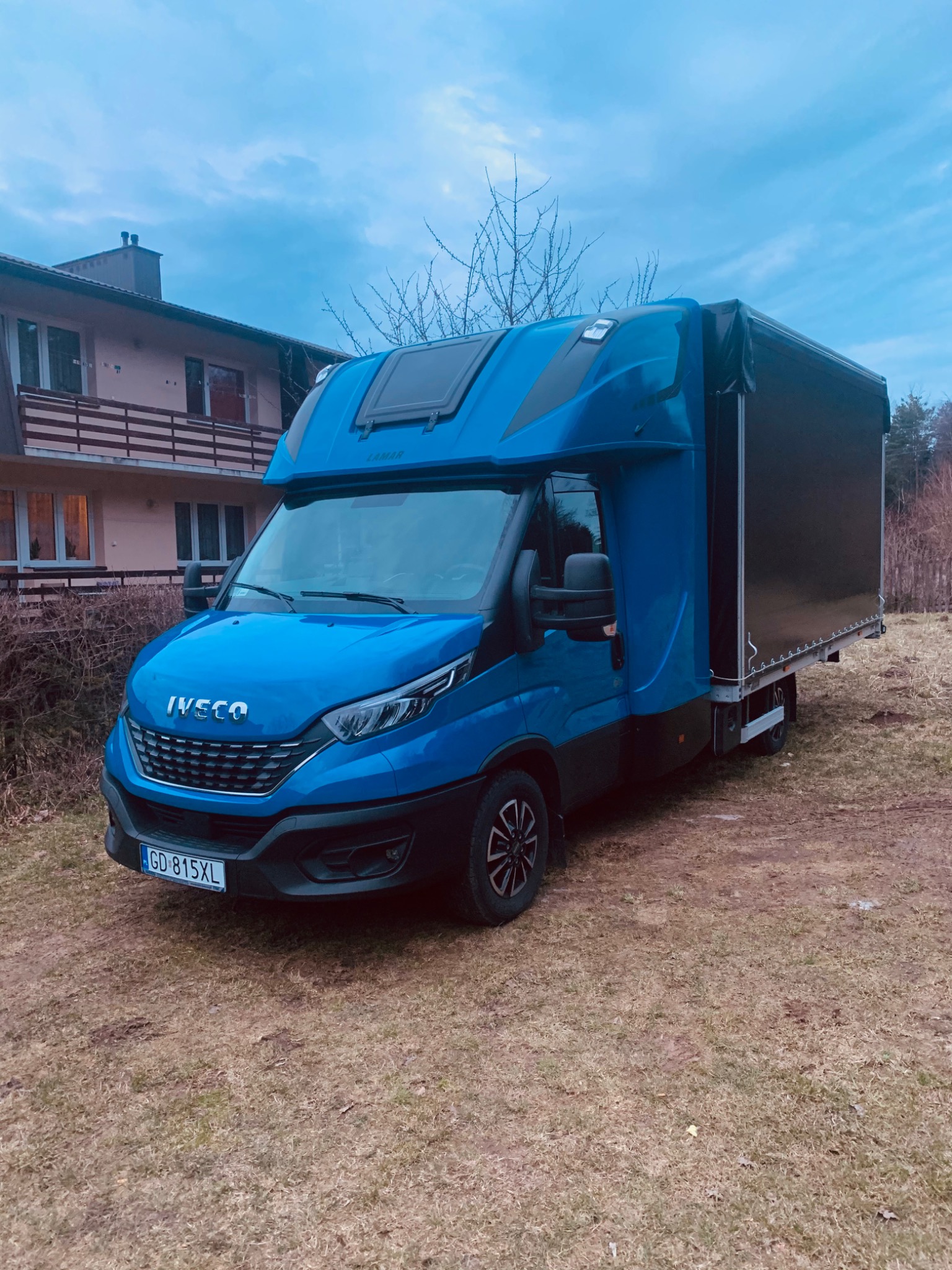 Niebieski samochód dostawczy Iveco z plandeką, zaparkowany na trawiastym terenie przed domem w pochmurny dzień.