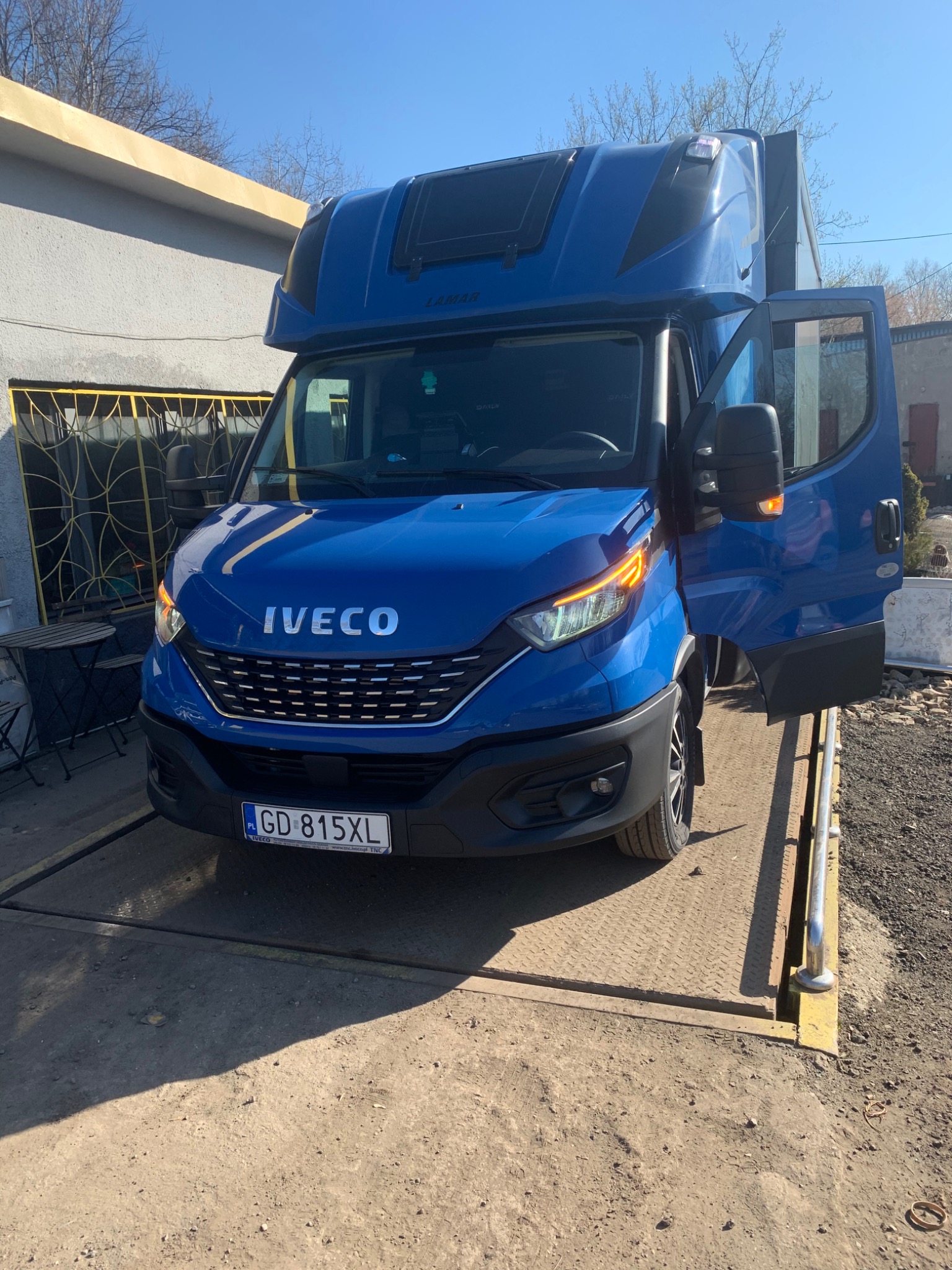 Niebieski samochód dostawczy Iveco z otwartymi drzwiami po stronie pasażera, zaparkowany na wadze samochodowej przed budynkiem.