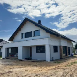 Nowoczesny dom jednorodzinny z jasną elewacją w trakcie wykończenia, widoczny fragment drewnianej okładziny, teren wokół domu częściowo przygotowany, niebo z chmurami.