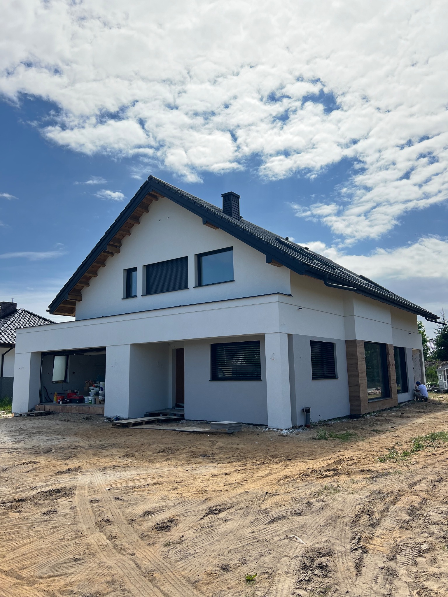 Nowoczesny dom jednorodzinny z jasną elewacją w trakcie wykończenia, widoczny fragment drewnianej okładziny, teren wokół domu częściowo przygotowany, niebo z chmurami.
