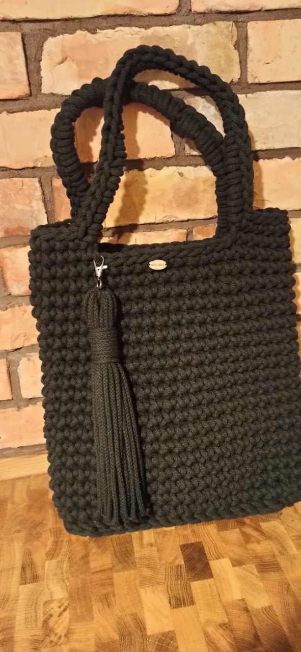 Czarna torebka wykonana na szydełku ze sznurka bawełnianego 5mm, z ozdobnym chwostem, prezentowana na tle ceglanej ściany i drewnianego blatu.