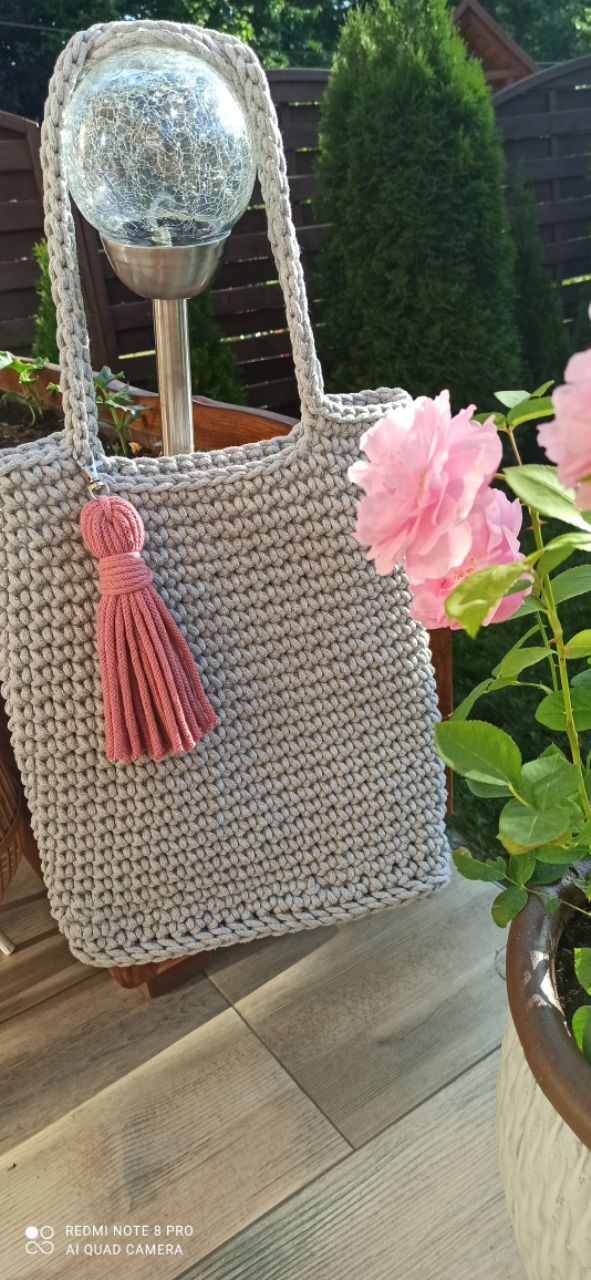 Szara torebka wykonana na szydełku ze sznurka bawełnianego 5mm, ozdobiona różowym chwostem, ustawiona na drewnianym krześle obok kwitnącego krzewu róży.