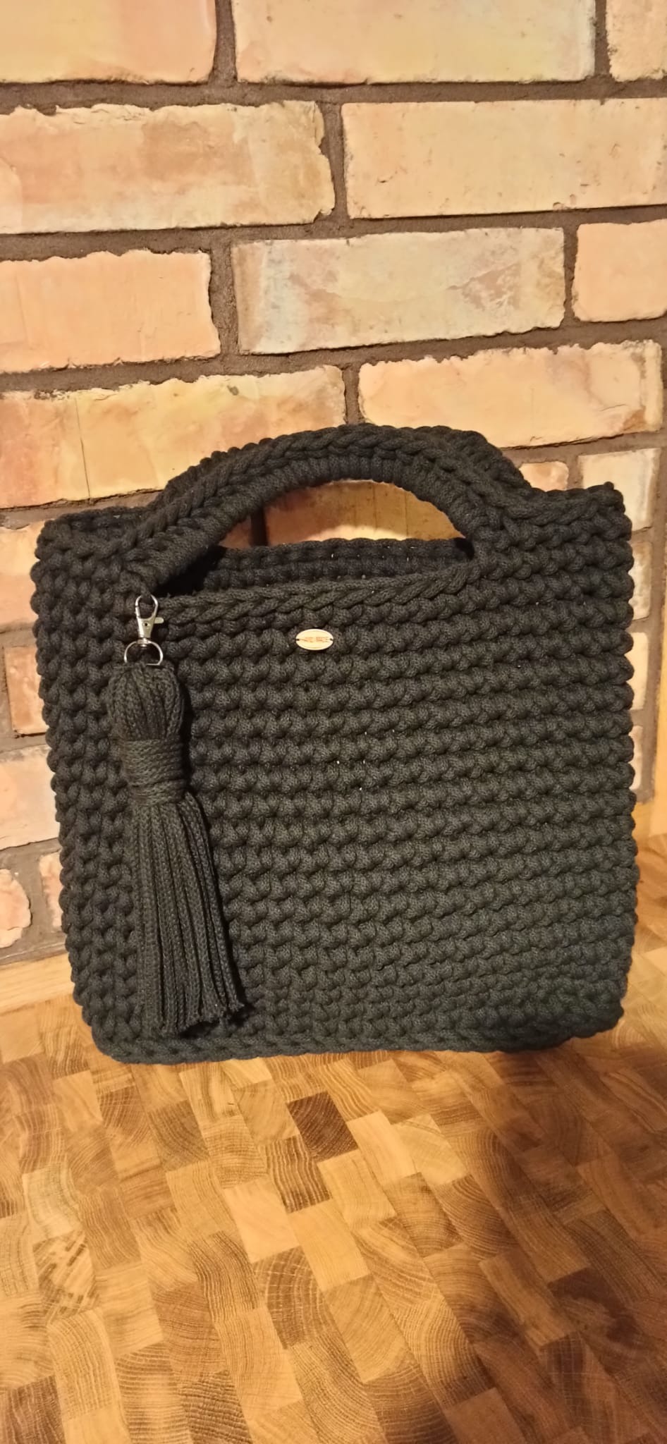 Ciemnoszara torebka damska, wykonana na szydełku, z grubego sznurka, z krótkimi uchwytami i ozdobnym chwostem, umieszczona na drewnianej podłodze na tle ceglanej ściany.