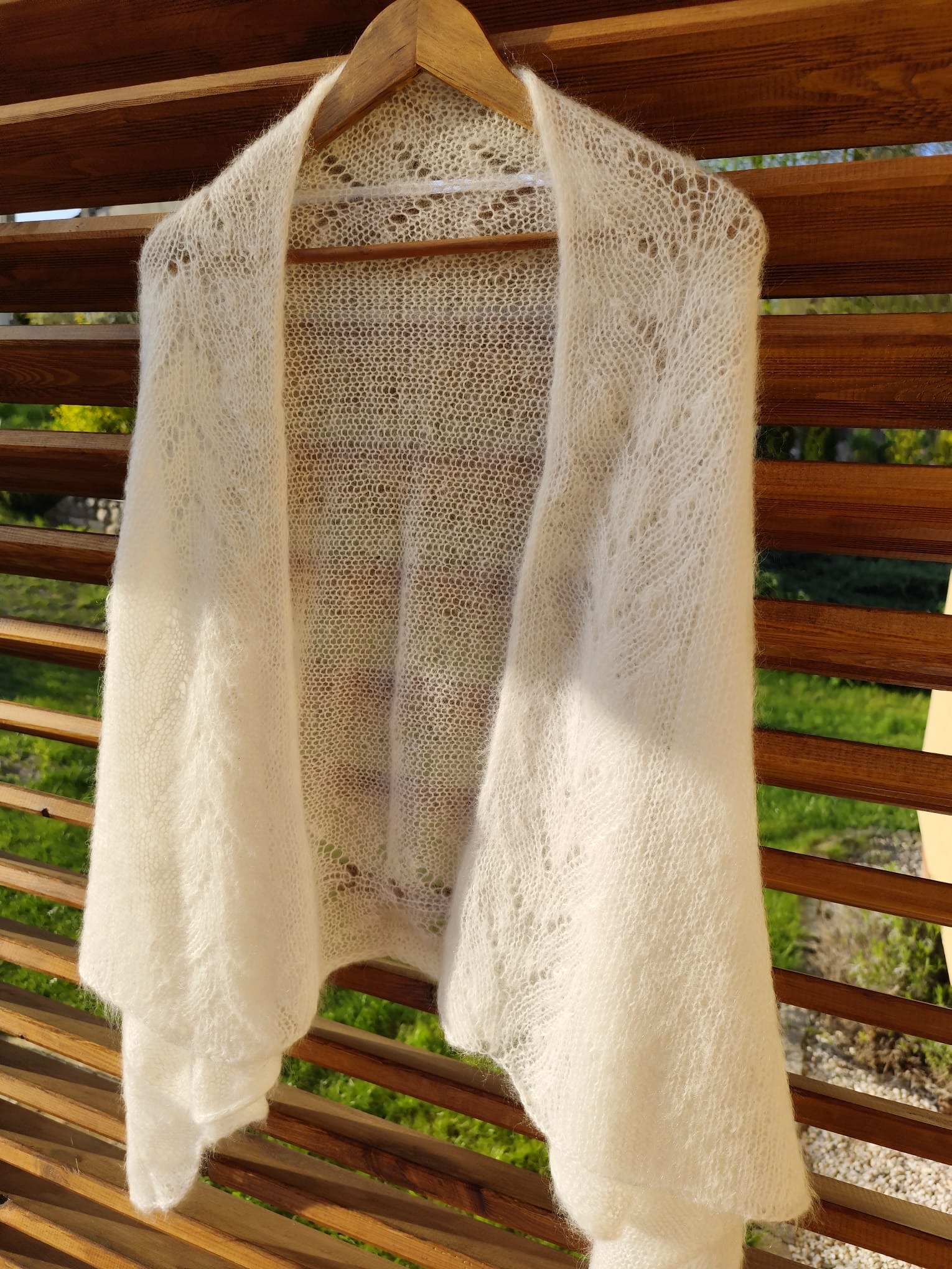 Delikatny, ażurowy, biały szal z moheru, zawieszony na drewnianym wieszaku na tle drewnianej konstrukcji i zieleni