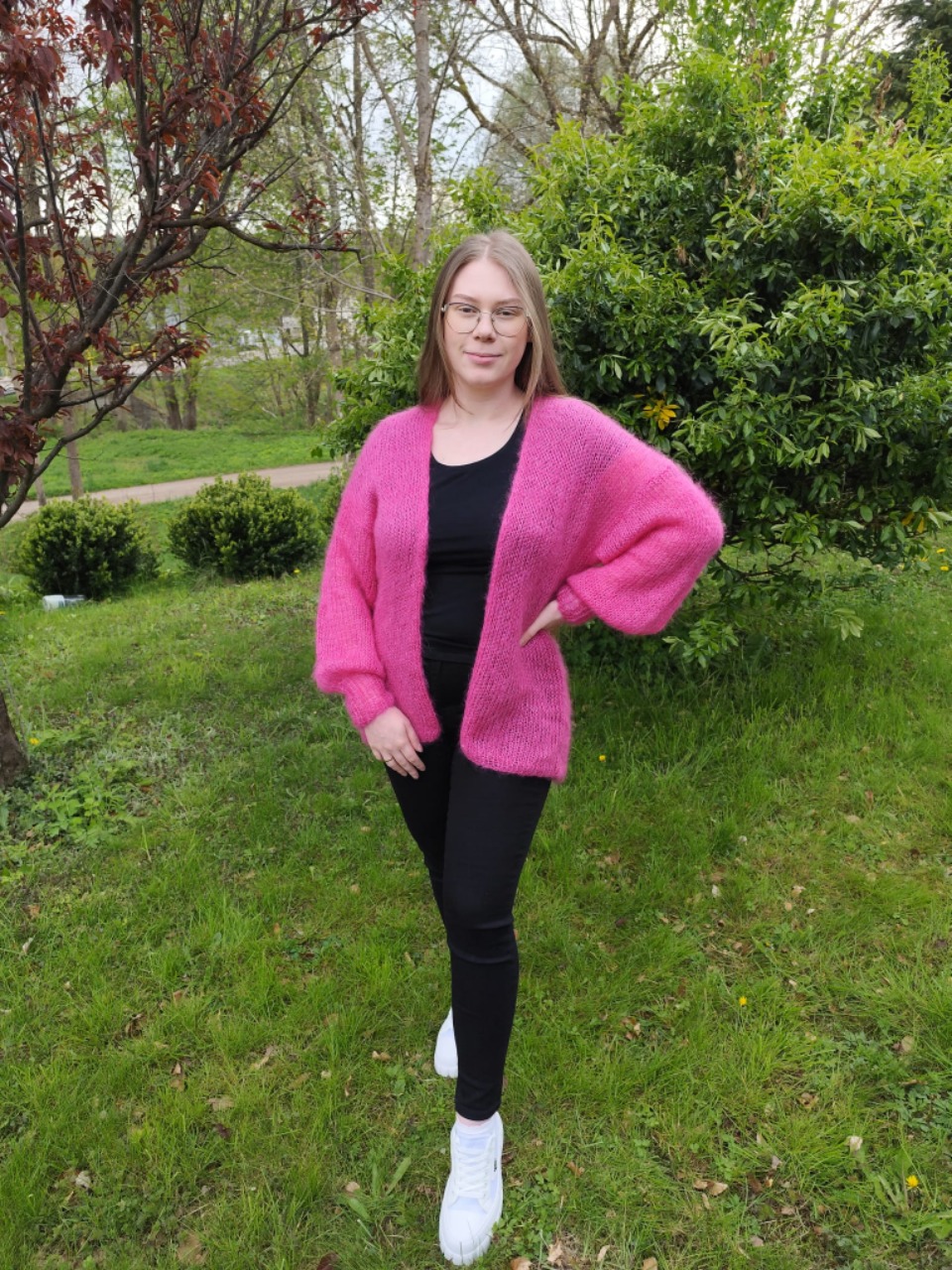 Kobieta w okularach prezentuje ręcznie robiony, puszysty, różowy kardigan z szerokimi rękawami na tle zielonego trawnika.