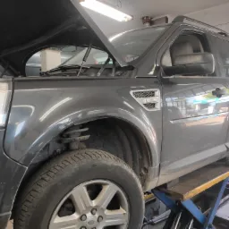 Ciemnoszary Land Rover na podnośniku warsztatowym z otwartą maską, brak koła, widoczne zawieszenie i opona z bieżnikiem.