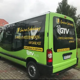 Zielony bus z naklejkami Gmina Łubniany i logo GTV BUS, zaparkowany na brukowanej powierzchni w pochmurny dzień.