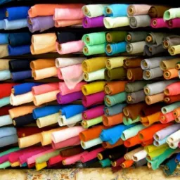 Regał pełen kolorowych, zwiniętych rolek tkanin w hurtowni, ułożonych warstwami, z widocznym wzorem na podłodze.