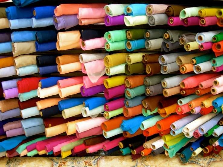 Regał pełen kolorowych, zwiniętych rolek tkanin w hurtowni, ułożonych warstwami, z widocznym wzorem na podłodze.