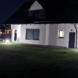 Nowoczesny dom jednorodzinny oświetlony kulistymi lampami ogrodowymi, widok nocny na elewację z czarnymi oknami i drzwiami.