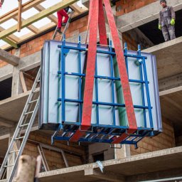 PM Technika Okienna - Montaż okien w surowym budynku: pakiet szyb z ramą transportowany dźwigiem, pracownicy na rusztowaniu i drabinie w tle.