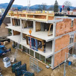 Montaż stolarki okiennej w budynku w trakcie budowy, widoczny dźwig, pracownicy i przygotowane ramy okienne.