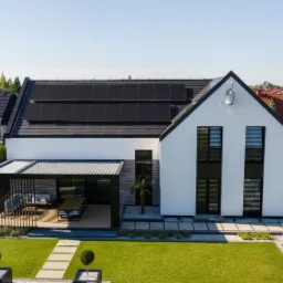 Nowoczesny dom jednorodzinny z czarnymi panelami słonecznymi na dachu, czarną pergolą nad tarasem i starannie utrzymanym trawnikiem z ozdobnymi krzewami.