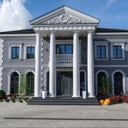 PM Technika Okienna - Elewacja okazałego, szarego domu z kolumnami, balkonem i symetrycznie rozmieszczonymi oknami, podkreślona jesiennymi dekoracjami z dyń.