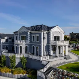 Dwupiętrowy dom w odcieniach szarości z białymi detalami architektonicznymi, balkonami z balustradami, ciemnym dachem i starannie zaprojektowanym terenem zielonym.