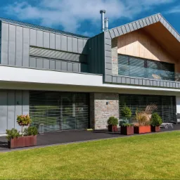 Nowoczesny dom z szarą blachą na elewacji i drewnianymi elementami, widoczne rolety zewnętrzne na oknach, taras z donicami i zielenią, zadbany trawnik.