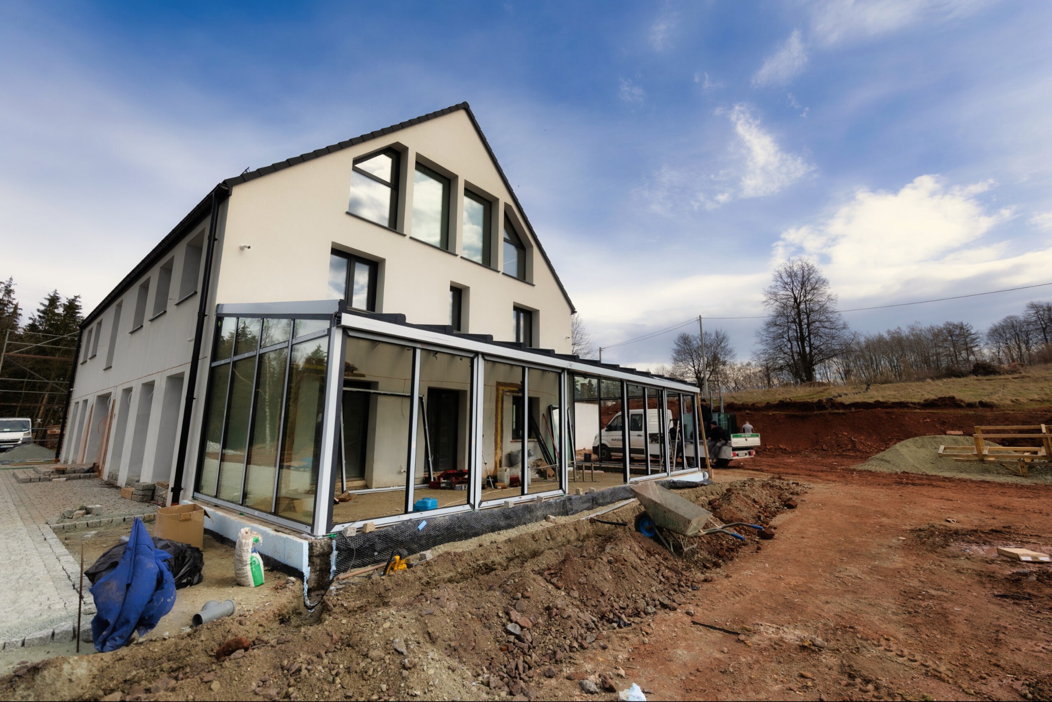 Budowa nowoczesnego domu z przeszkloną werandą w trakcie prac wykończeniowych, widoczne materiały budowlane i wykop pod fundamenty.