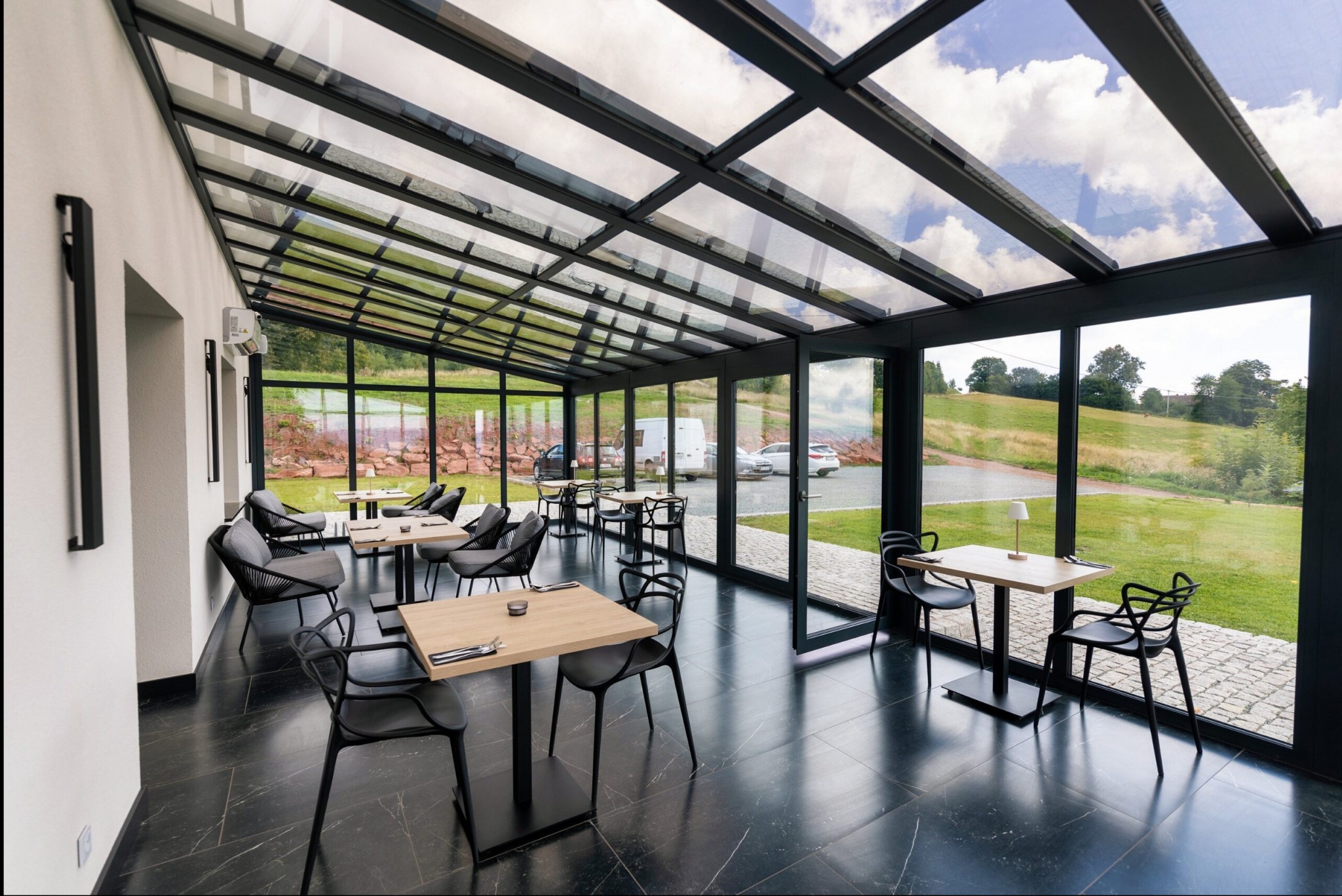 Szklana weranda z czarną ramą okienną w Wałbrzychu, widok na zewnątrz z zielonym wzgórzem i zaparkowanymi samochodami, czarne krzesła i stoliki wewnątrz.