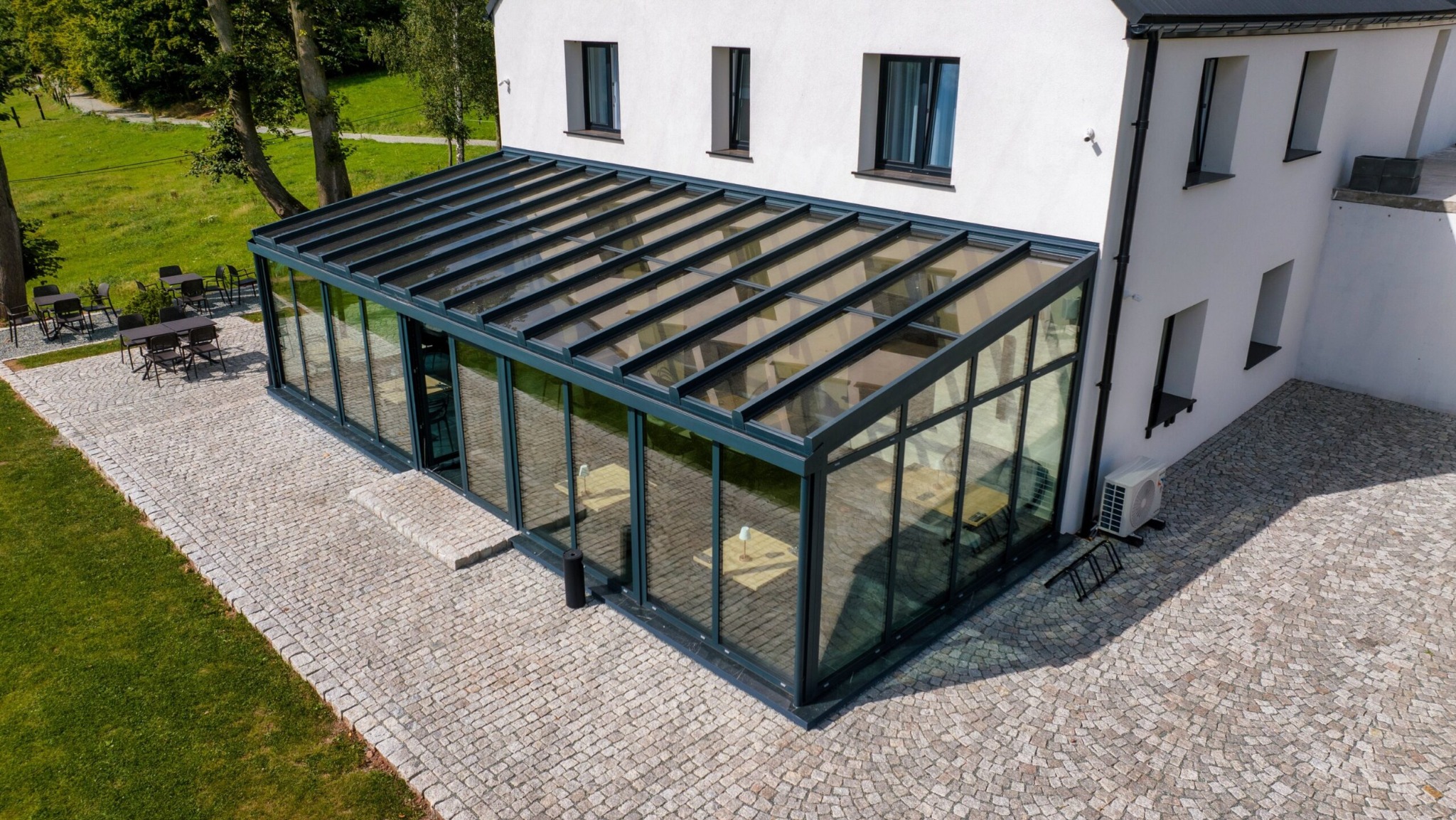 Szklana oranżeria z ciemną ramą dobudowana do białego budynku, widok z góry na kamienny bruk i trawnik.