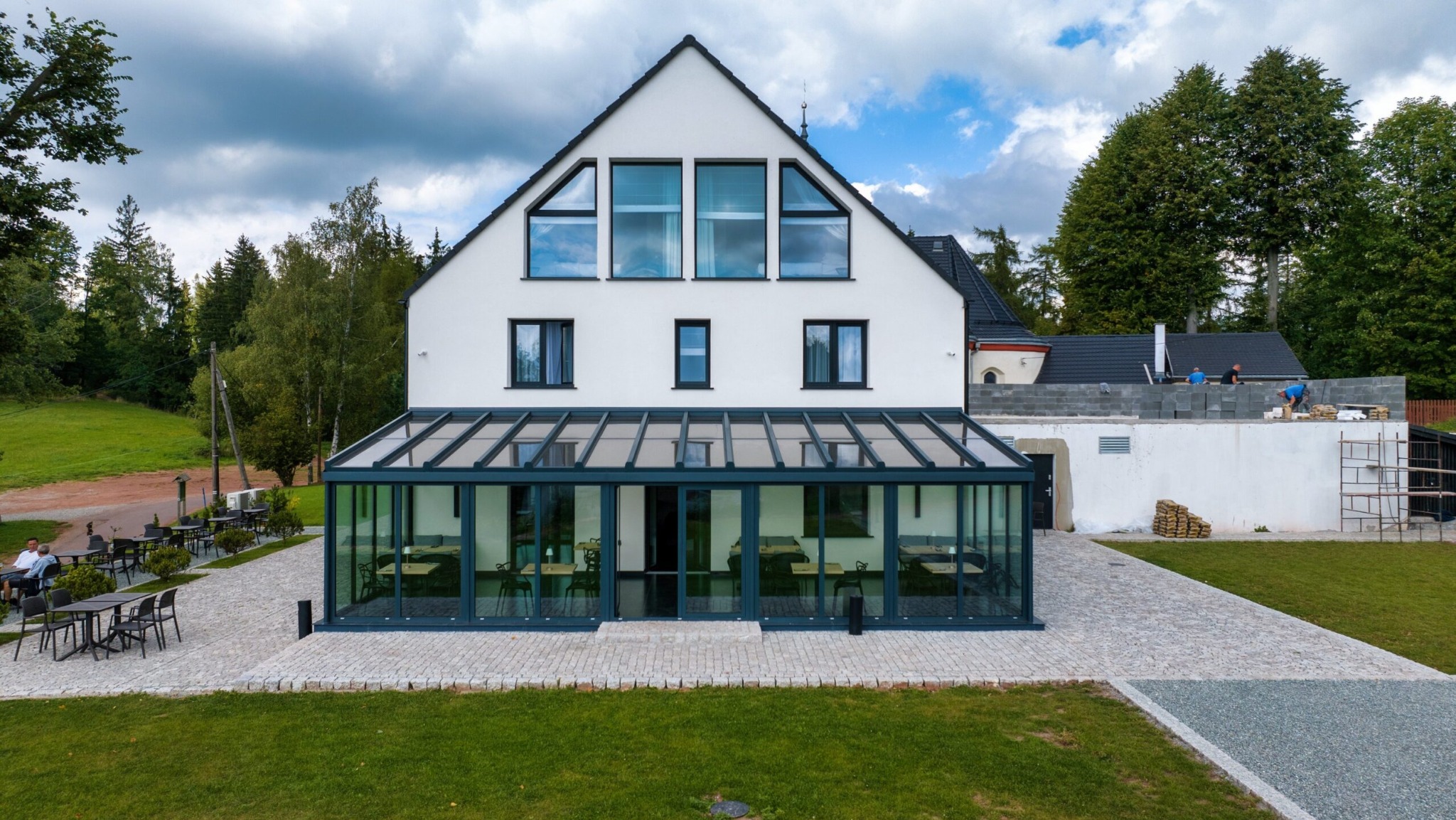 Nowoczesny dom z przeszkloną werandą w stylu ogrodu zimowego, widoczne ciemne ramy okien i drzwi, otoczenie zieleni i plac z kostki brukowej.