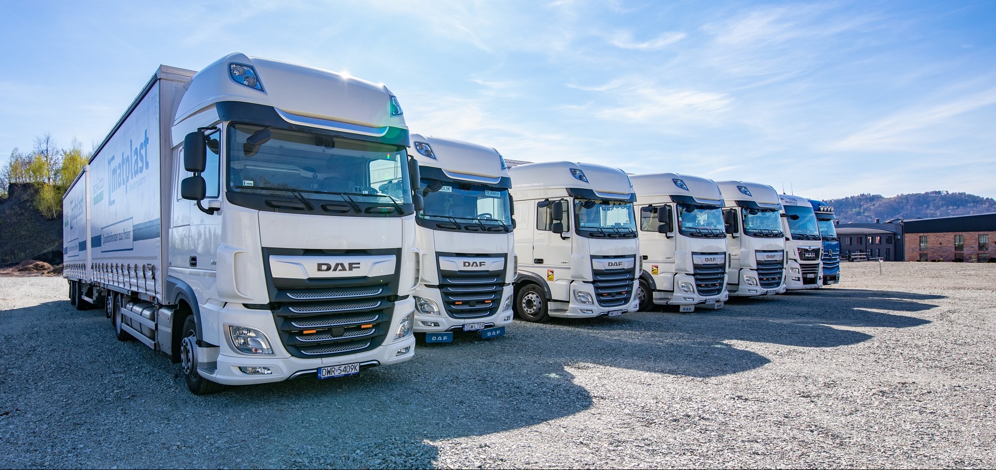 Rząd białych ciężarówek DAF i MAN zaparkowanych na żwirowym placu w słoneczny dzień, z budynkiem w tle.