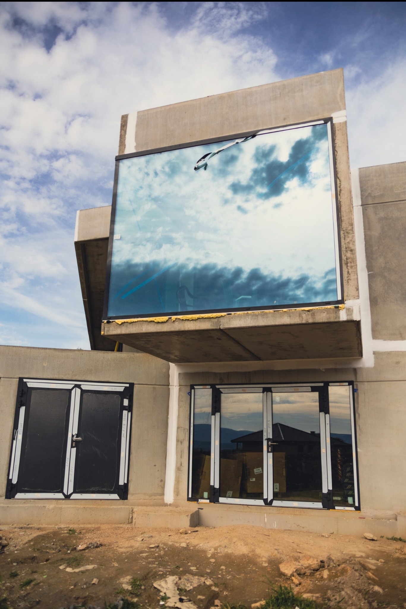 Surowy stan budynku w trakcie montażu stolarki okiennej: duże, czarne ramy okienne i drzwiowe z folią ochronną, częściowo odbijające niebo, osadzone w betonowej konstrukcji.