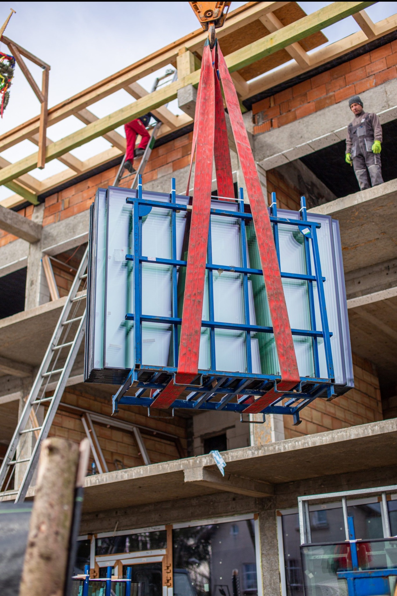 Montaż okien w surowym budynku: pakiet szyb z ramą transportowany dźwigiem, pracownicy na rusztowaniu i drabinie w tle.