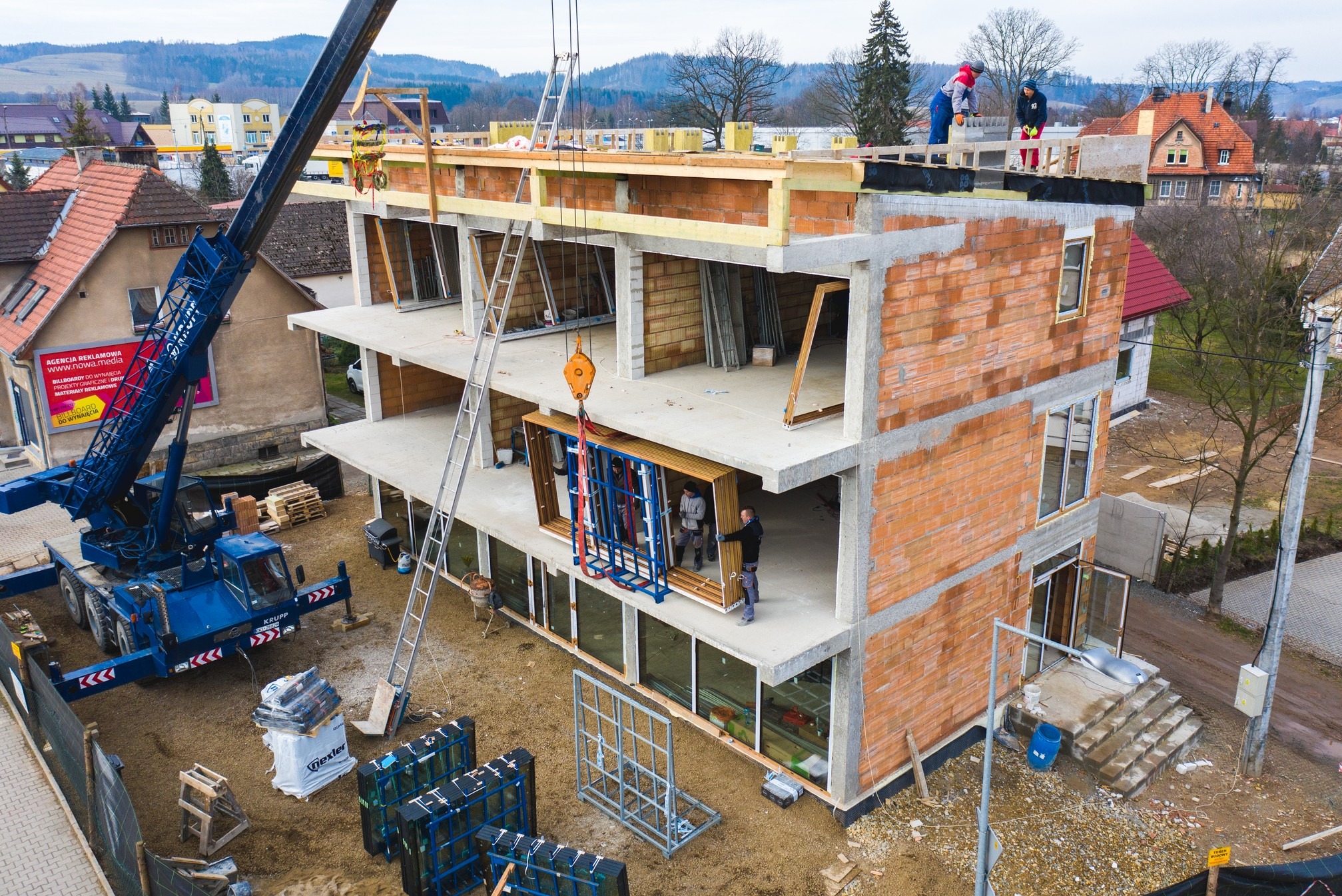 Montaż stolarki okiennej w budynku w trakcie budowy, widoczny dźwig, pracownicy i przygotowane ramy okienne.