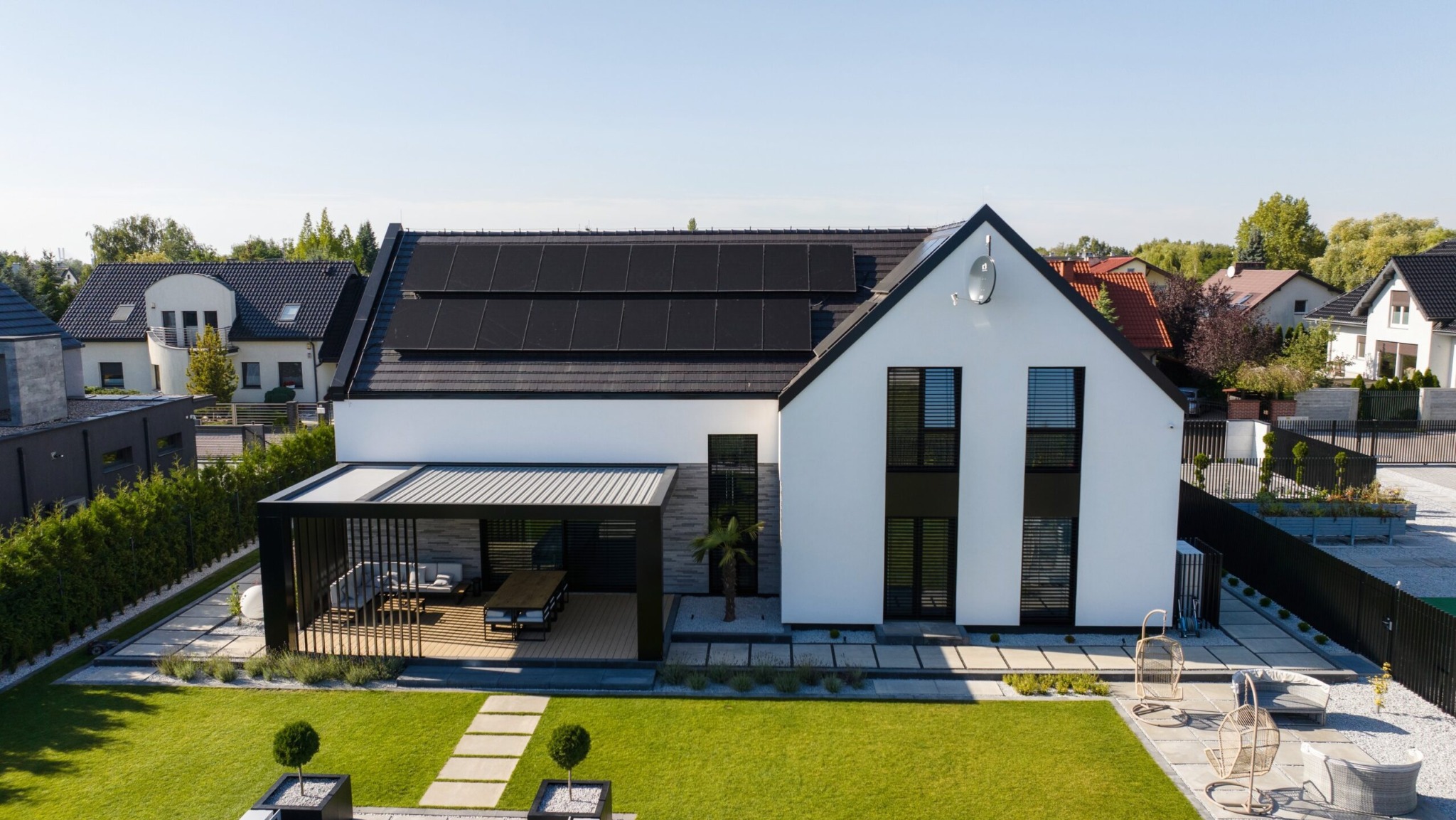 Nowoczesny dom jednorodzinny z czarnymi panelami słonecznymi na dachu, czarną pergolą nad tarasem i starannie utrzymanym trawnikiem z ozdobnymi krzewami.