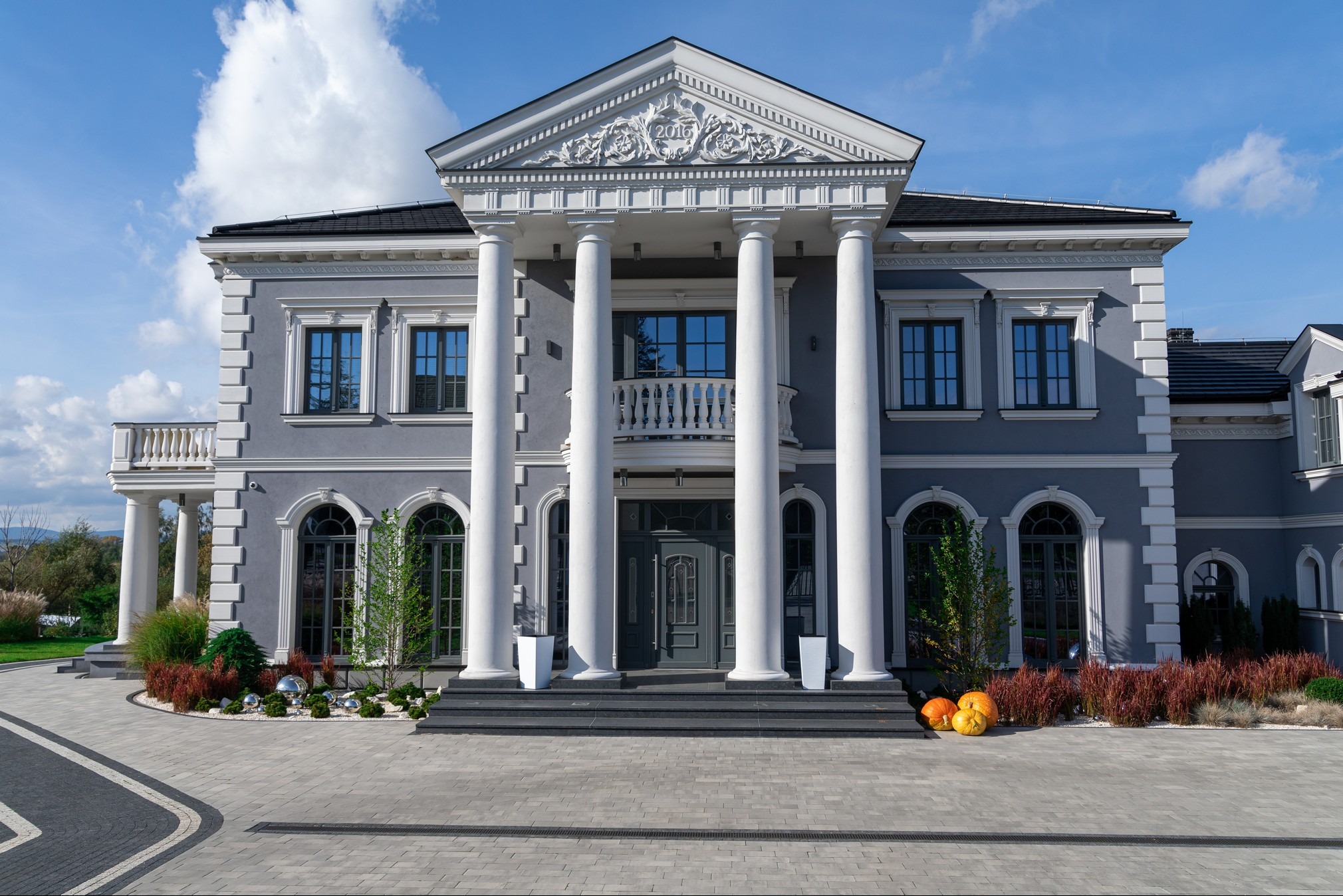 Elewacja okazałego, szarego domu z kolumnami, balkonem i symetrycznie rozmieszczonymi oknami, podkreślona jesiennymi dekoracjami z dyń.