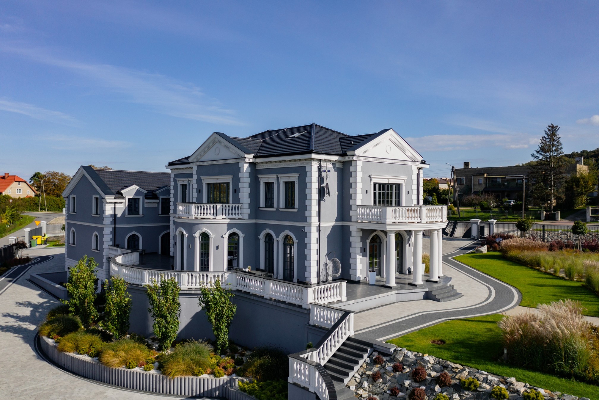 Dwupiętrowy dom w odcieniach szarości z białymi detalami architektonicznymi, balkonami z balustradami, ciemnym dachem i starannie zaprojektowanym terenem zielonym.