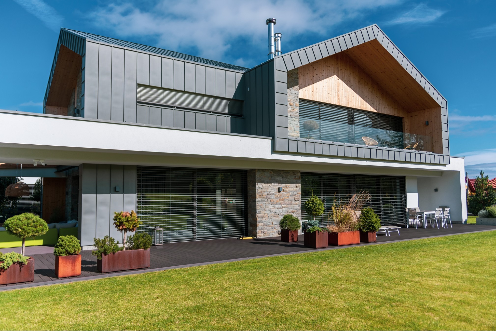 Nowoczesny dom z szarą blachą na elewacji i drewnianymi elementami, widoczne rolety zewnętrzne na oknach, taras z donicami i zielenią, zadbany trawnik.