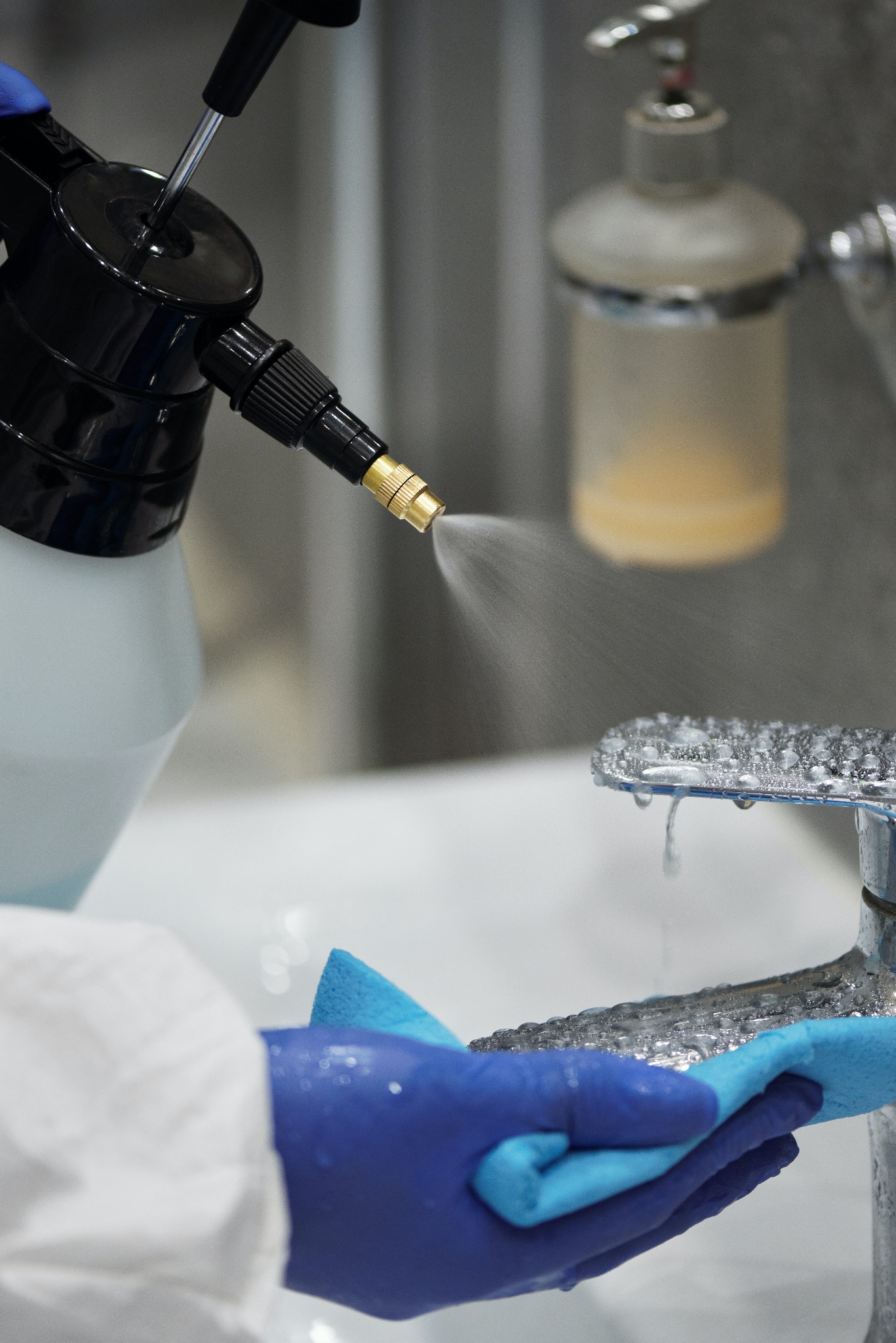 Czyszczenie baterii umywalkowej środkiem dezynfekującym za pomocą spryskiwacza i ściereczki z mikrofibry, osoba w rękawicach ochronnych.