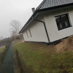 Świeżo otynkowany dom jednorodzinny z ciemną dachówką i grafitową stolarką okienną, widok z boku na tle pochmurnego nieba i zieleni, z widoczną siatką ogrodzeniową w tle.