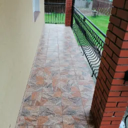 Wyłożony wzorzystymi płytkami taras z ceglanymi słupkami i czarną, metalową balustradą, widok na zielony ogród.