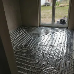 Instalacja ogrzewania podłogowego w pomieszczeniu z widokiem na zewnątrz przez okno, rury ułożone na folii izolacyjnej i siatce montażowej.