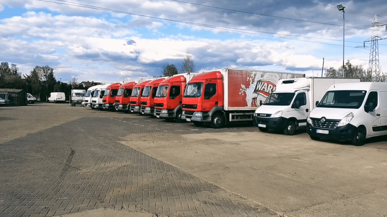 Rząd czerwonych ciężarówek i białych busów zaparkowanych na brukowanym placu pod pochmurnym niebem, widoczna reklama piwa na jednej z ciężarówek.