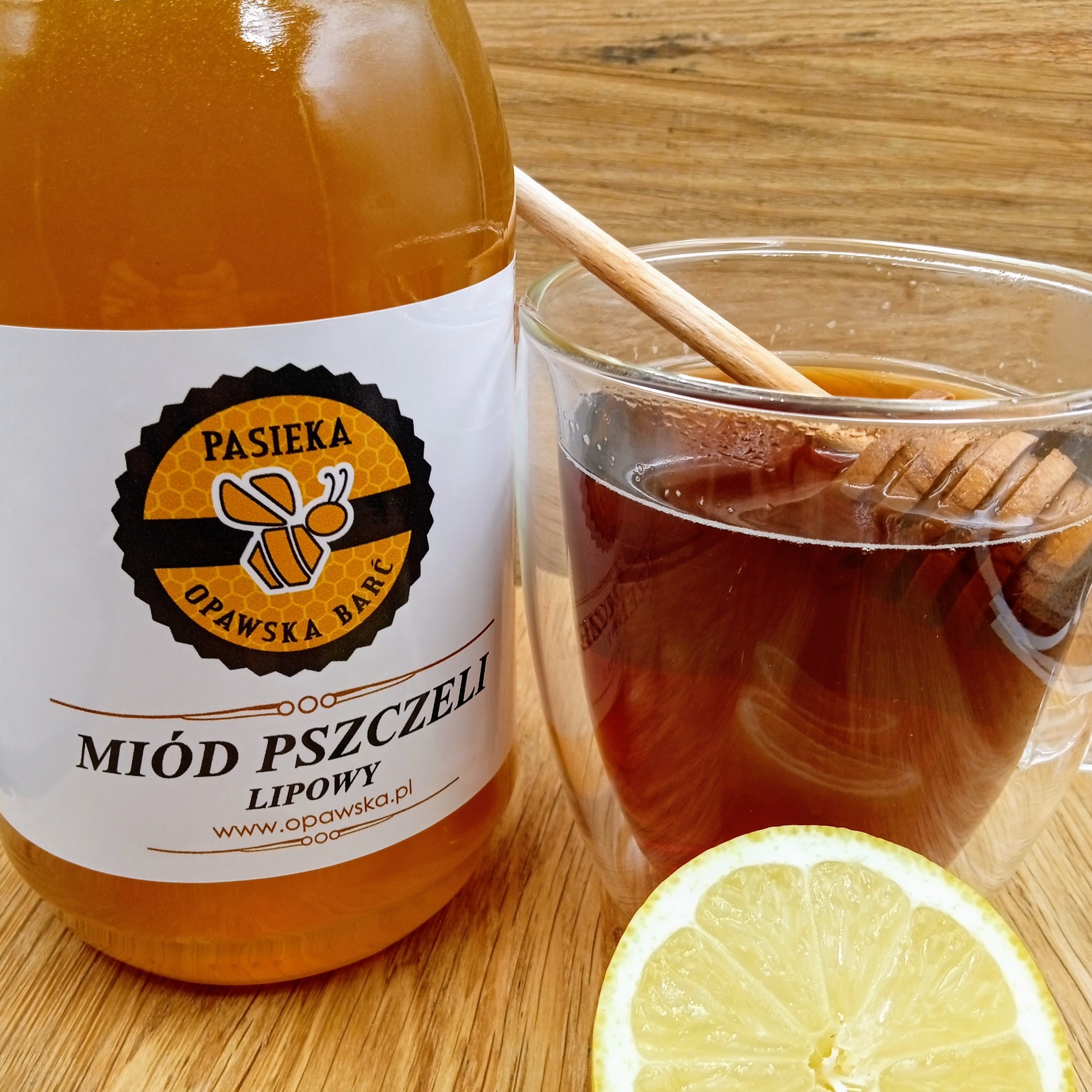Butelka z etykietą Pasieka Opawska Barć, szklanka herbaty z drewnianym nabierakiem i plaster cytryny na drewnianym blacie.