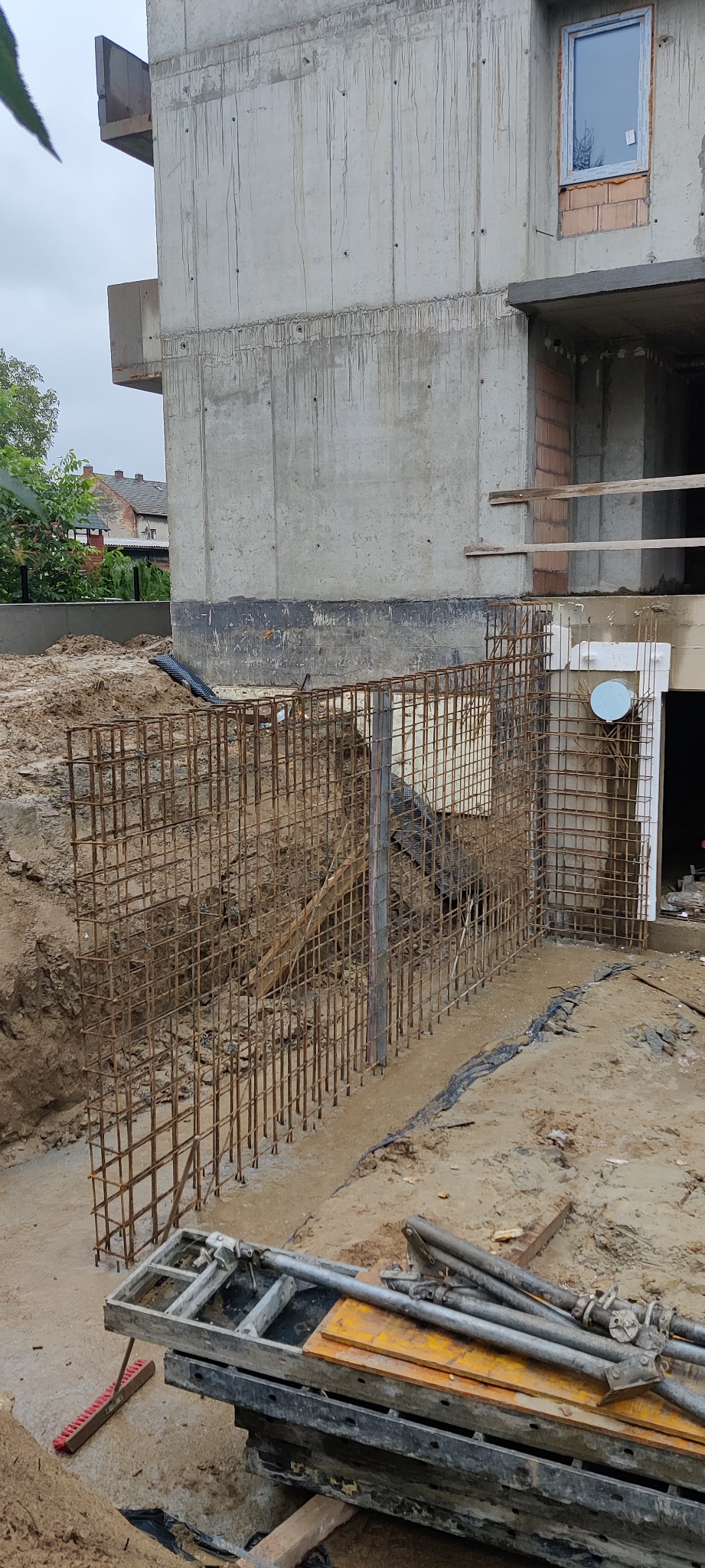 Zbrojenie pionowe fundamentów budynku w trakcie budowy, widoczne pręty stalowe i szalunki, w tle surowy beton konstrukcji i wykop ziemny.