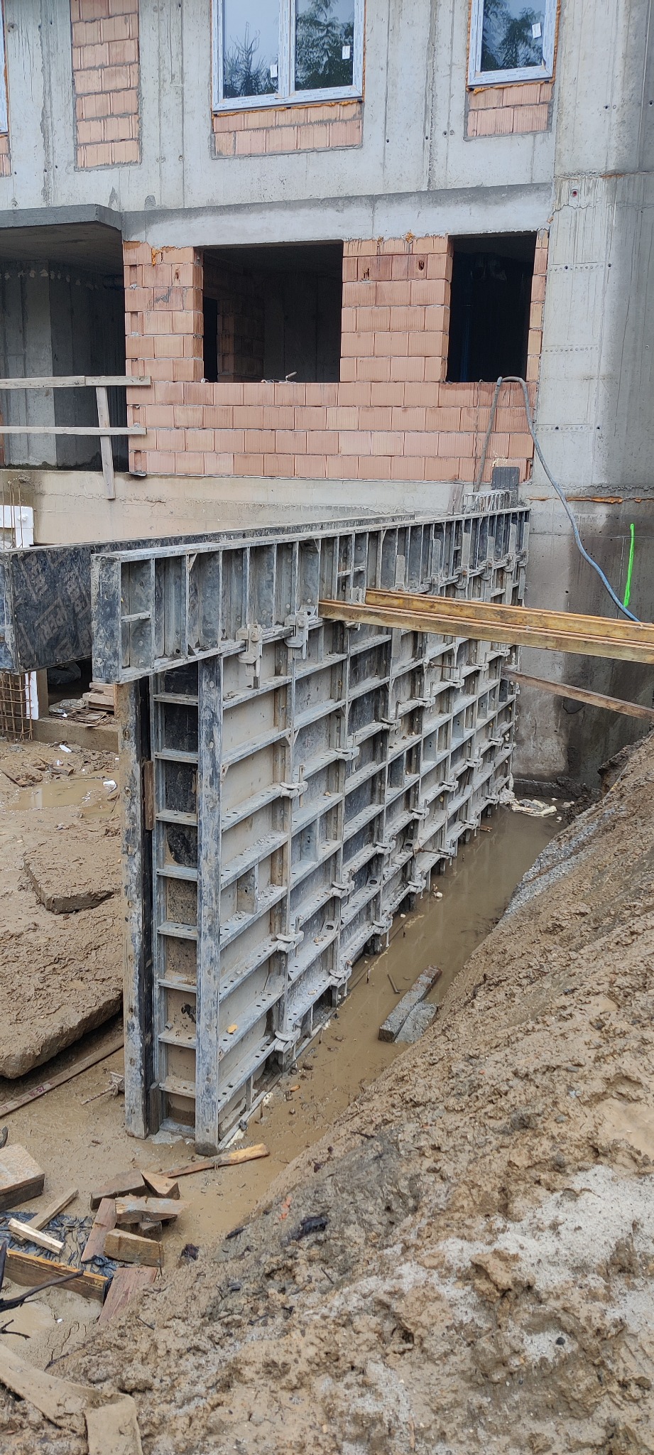 Szare szalunki fundamentowe w wykopie wypełnionym błotnistą wodą, z fragmentem budynku w trakcie budowy w tle.