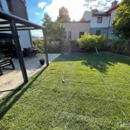 Nowoczesny taras z zadaszeniem przy domu, przylegający do zadbanego trawnika z widocznym wężem ogrodowym w słoneczny dzień.