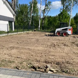 Przygotowany teren pod trawnik lub ogród z użyciem miniładowarki Bobcat S100, widoczna świeżo wyrównana ziemia, obok nowoczesny dom z grafitowymi akcentami i ogrodzenie z siatki.