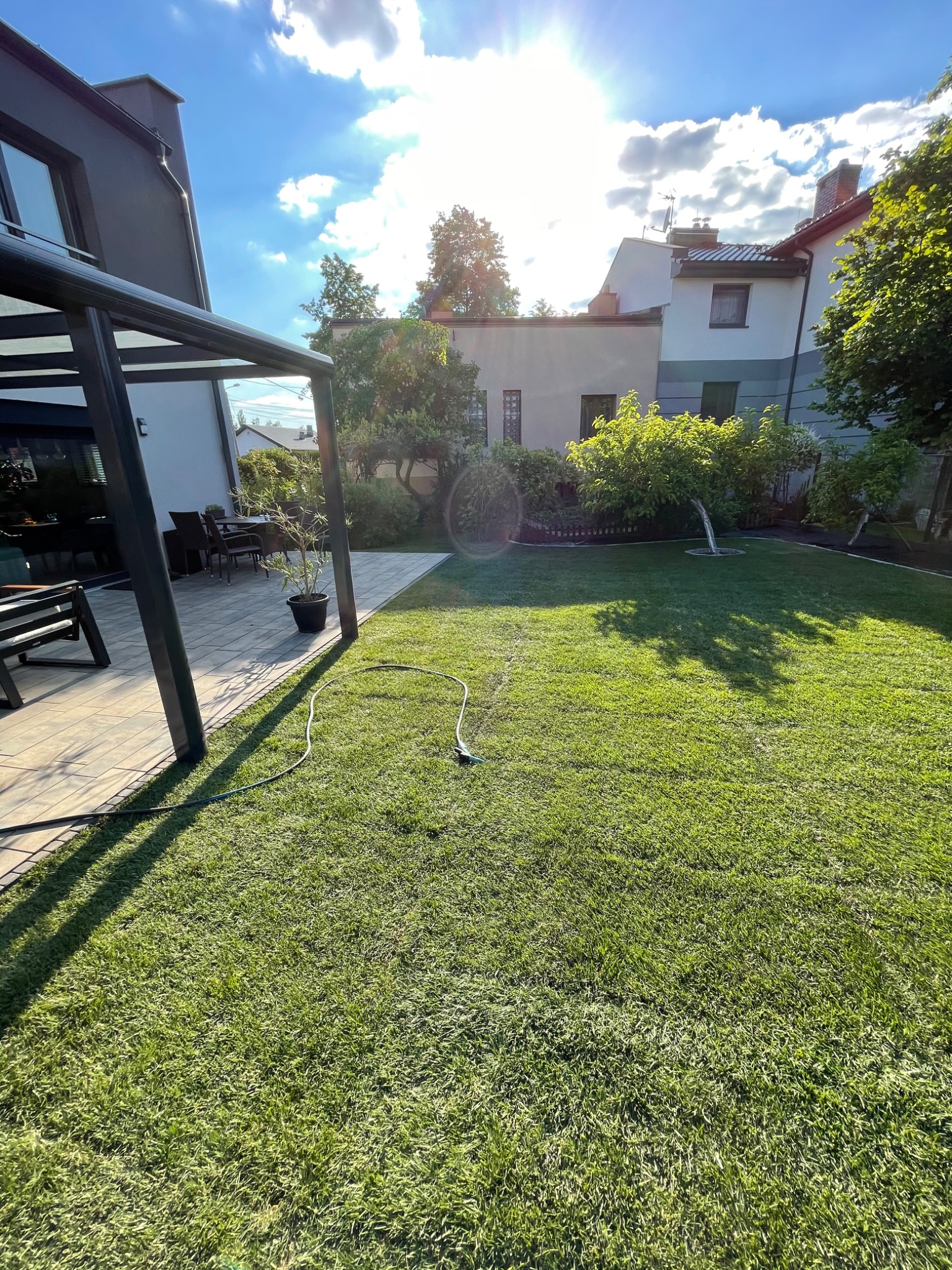 Nowoczesny taras z zadaszeniem przy domu, przylegający do zadbanego trawnika z widocznym wężem ogrodowym w słoneczny dzień.