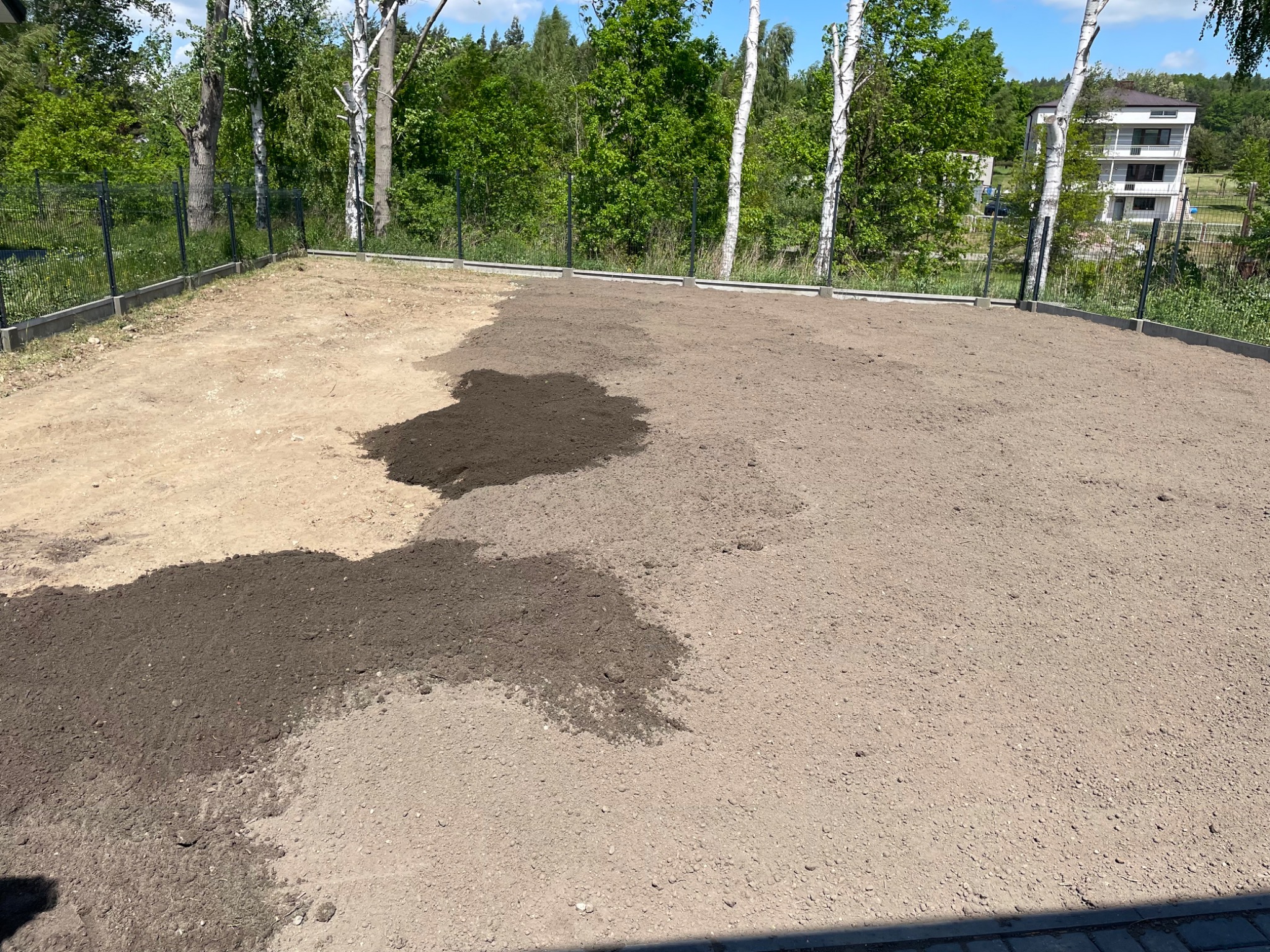 Przygotowanie terenu pod trawnik: wyrównana ziemia w ogrodzie, część powierzchni pokryta świeżą, ciemną ziemią, otoczenie z siatki i drzew.