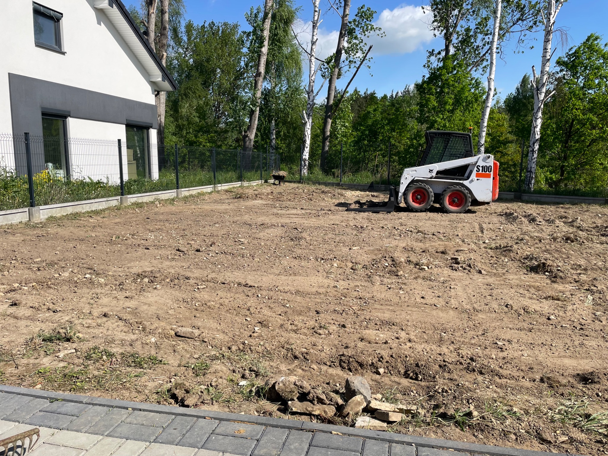 Przygotowany teren pod trawnik lub ogród z użyciem miniładowarki Bobcat S100, widoczna świeżo wyrównana ziemia, obok nowoczesny dom z grafitowymi akcentami i ogrodzenie z siatki.