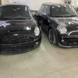 Dwa czarne Mini Cooper S stojące w warsztacie, jeden z widocznym otwartym szyberdachem, drugi z felgami aluminiowymi, widok z przodu.