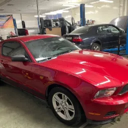 Czerwony Ford Mustang w warsztacie samochodowym, widoczny podnośnik i inne pojazdy w tle.