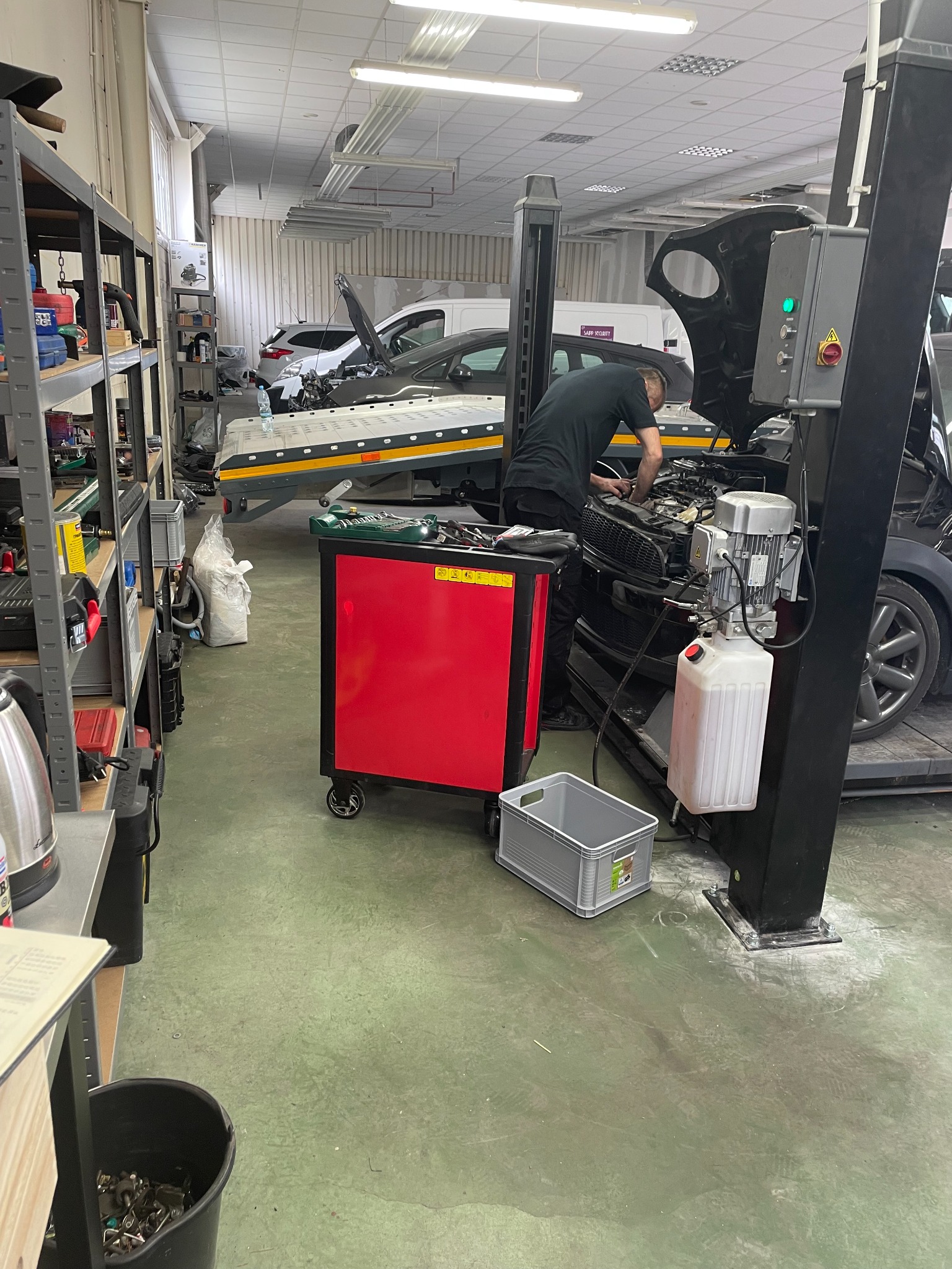 Warsztat samochodowy: Mężczyzna w czarnej koszulce pochyla się nad otwartą maską czarnego samochodu na podnośniku. W tle widoczna laweta z innym pojazdem. Czerwony wózek narzędziowy i półki...